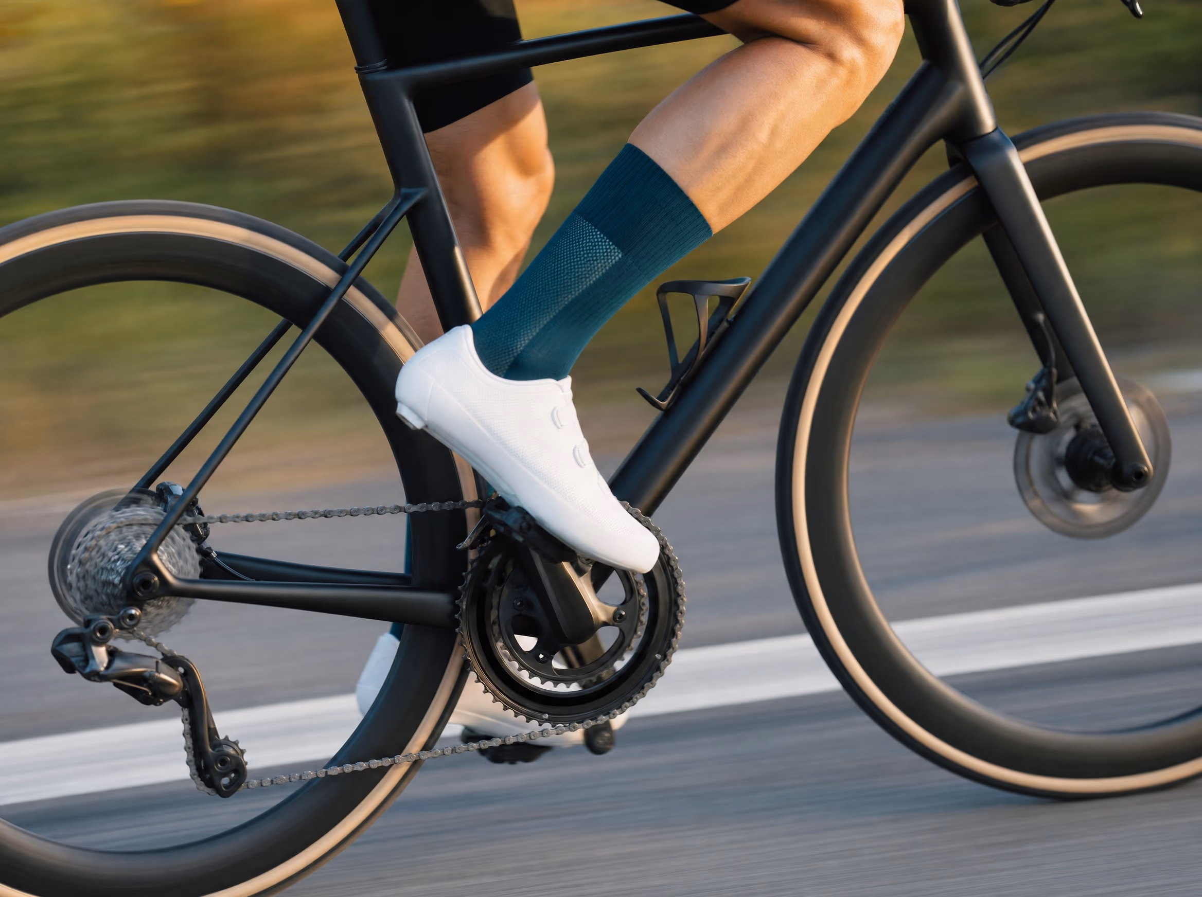 A road cyclist mid-pedal wearing a deep-teal performance cycling sock and white shoe, shot in motion on a sunlit road