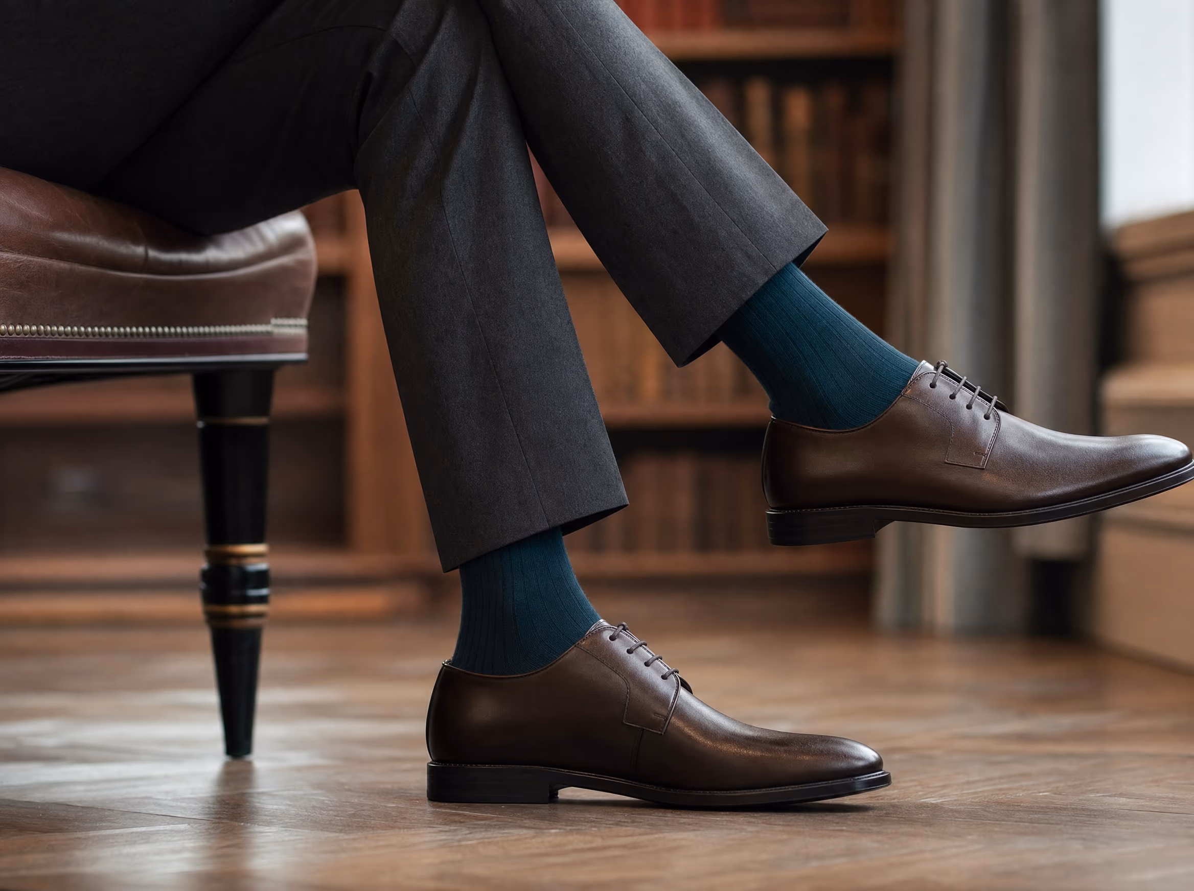 A seated man's crossed legs in tailored trousers, wearing a fine-gauge deep-teal ribbed dress sock above a polished brown leather derby shoe